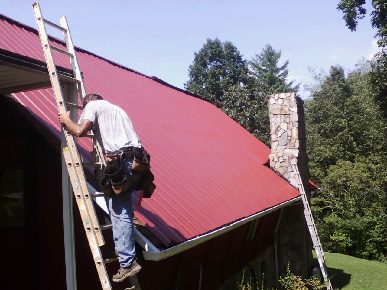 Roofer on a ladder beside a red metal roof and stone chimney — Soffit & Fascia Repair by Roofing Brothers in Haskins